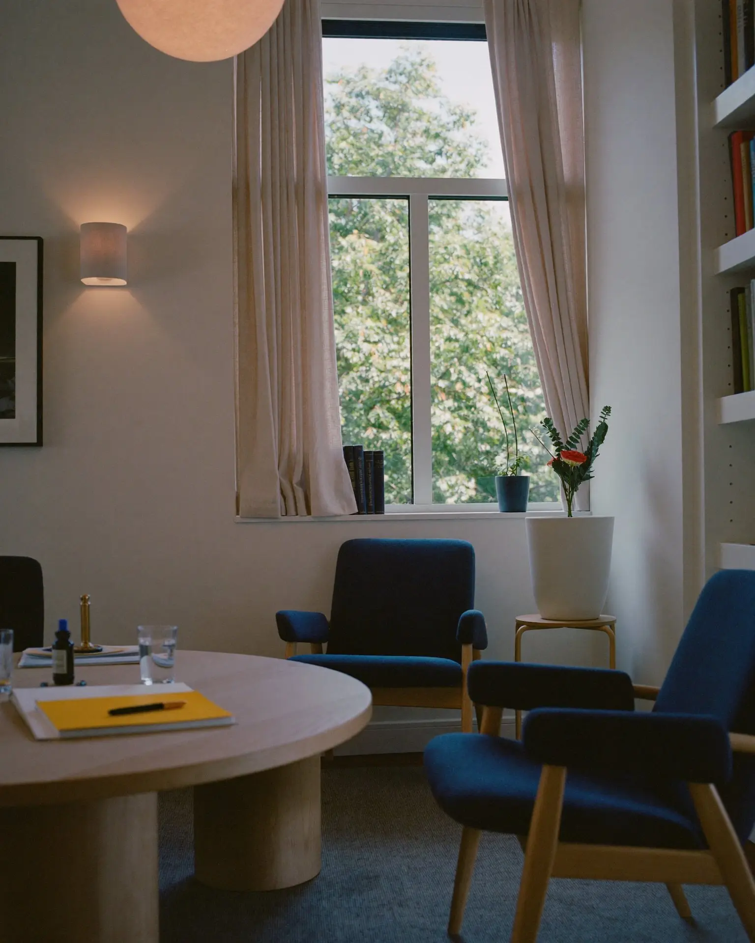 A bright consultation room with a round oak table, a yellow legal pad, two glasses of water and navy armchairs by a tall window